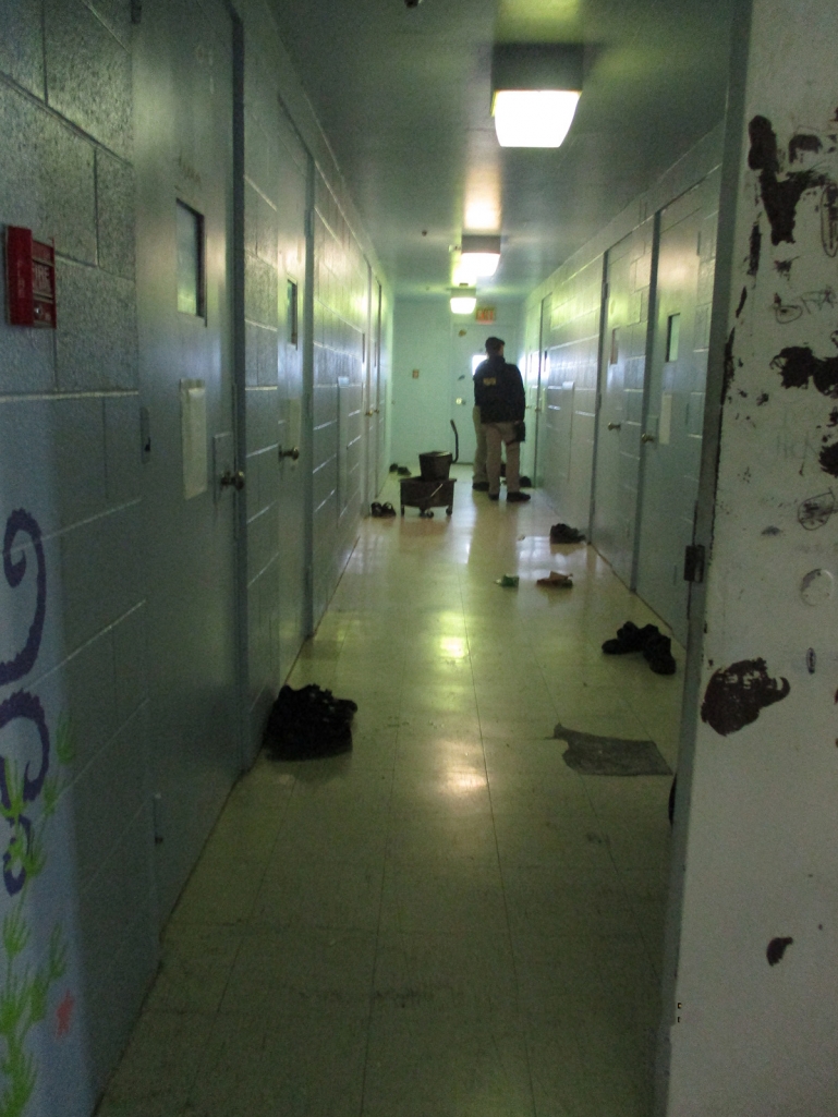 A hallway at KCJC with fluorescent lights, walls made of cinderblocks, and peeling linoleum floors. Detained youths’ shoes are placed outside each cell closed door and probation staff are standing at the end of the hallway.
