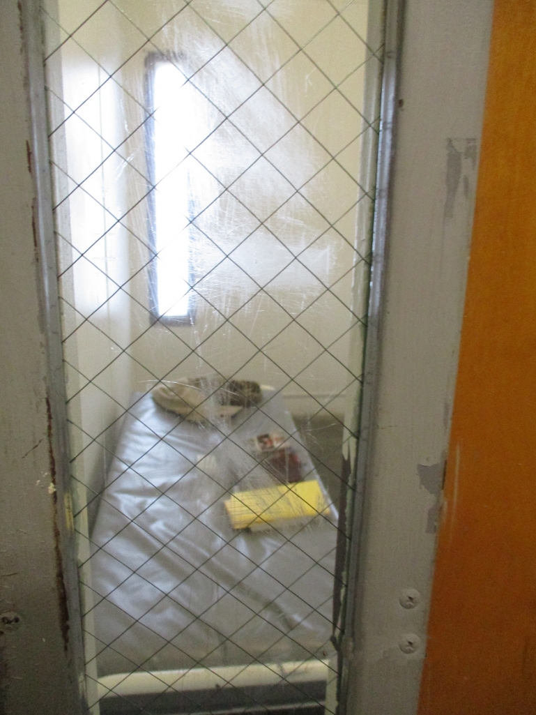 Another cell at KCJC as seen through the small window in the closed door. The mattress is bare except for some books, a notebook, and some clothes. The door’s window is scratched.