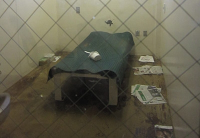 Inside of a dirty jail cell with garbage around bed.