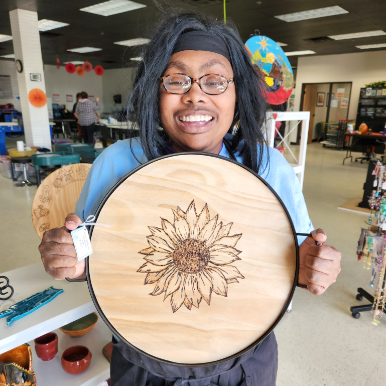 Jemel Williams holding one of her pieces titled 'Sunflower'. A picture of Jemel Williams smiling while holding one of her works of art titled 'Sunflower'. This work is a wood burned sunflower graphic on a circular piece of raw, light-colored, wood.