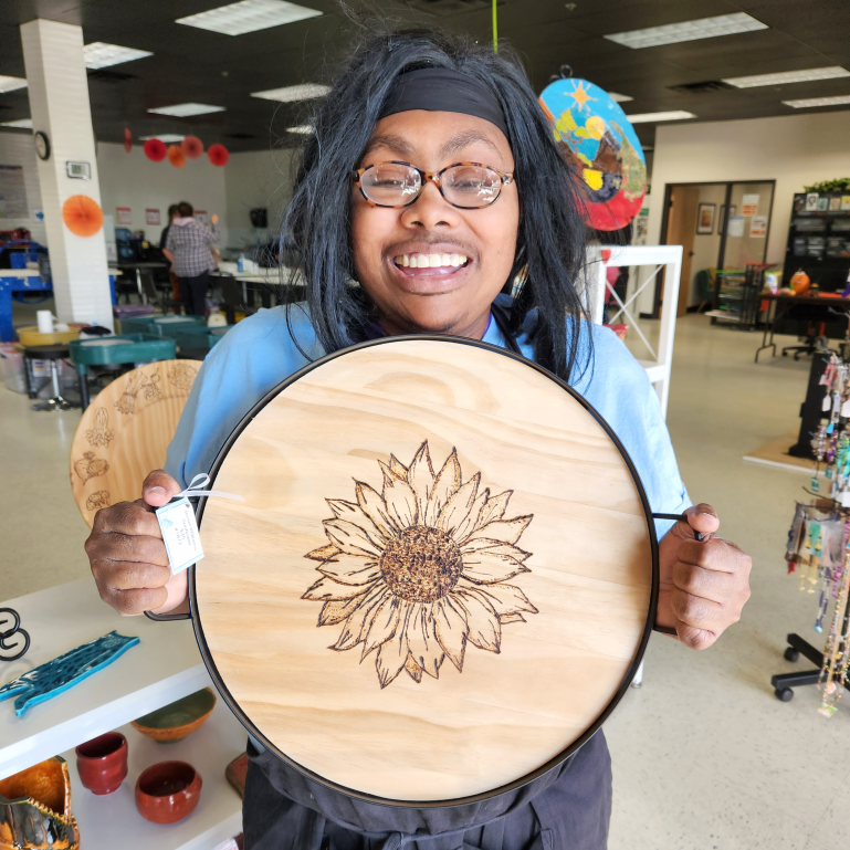 A picture of Jemel Williams smiling while holding one of her works of art titled 'Sunflower'. This work is a wood burned sunflower graphic on a circular piece of raw, light-colored, wood.