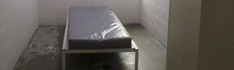 A cell at KCJC. The room has concrete floors and cinderblock walls, and is empty except for a bare mattress on a bed frame bolted to the floor and a metal toilet.