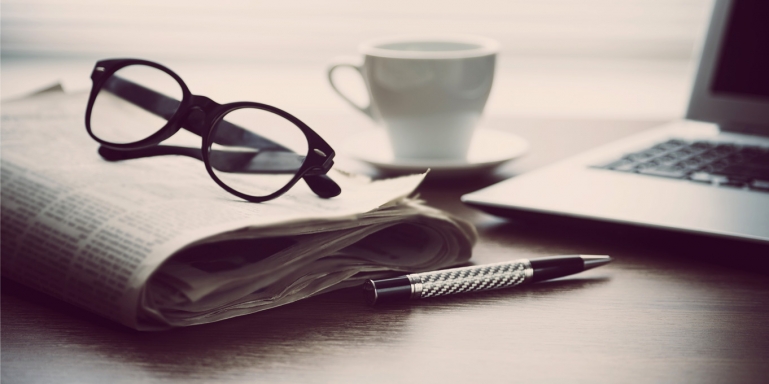 Image of a folder newspaper, eye glasses and a laptop sitting on a desk.