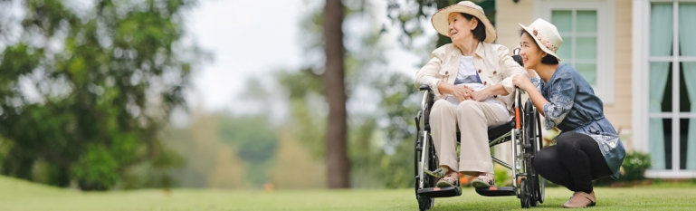 Photo of a elderly woman in a wheelchair with her daughter kneeling next to her. They are in their backyard on a beautiful day.