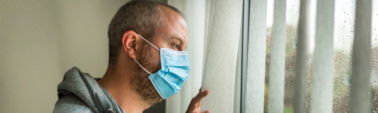 Photo of a man with a medical mask on looking out a window. He is clearly depressed.