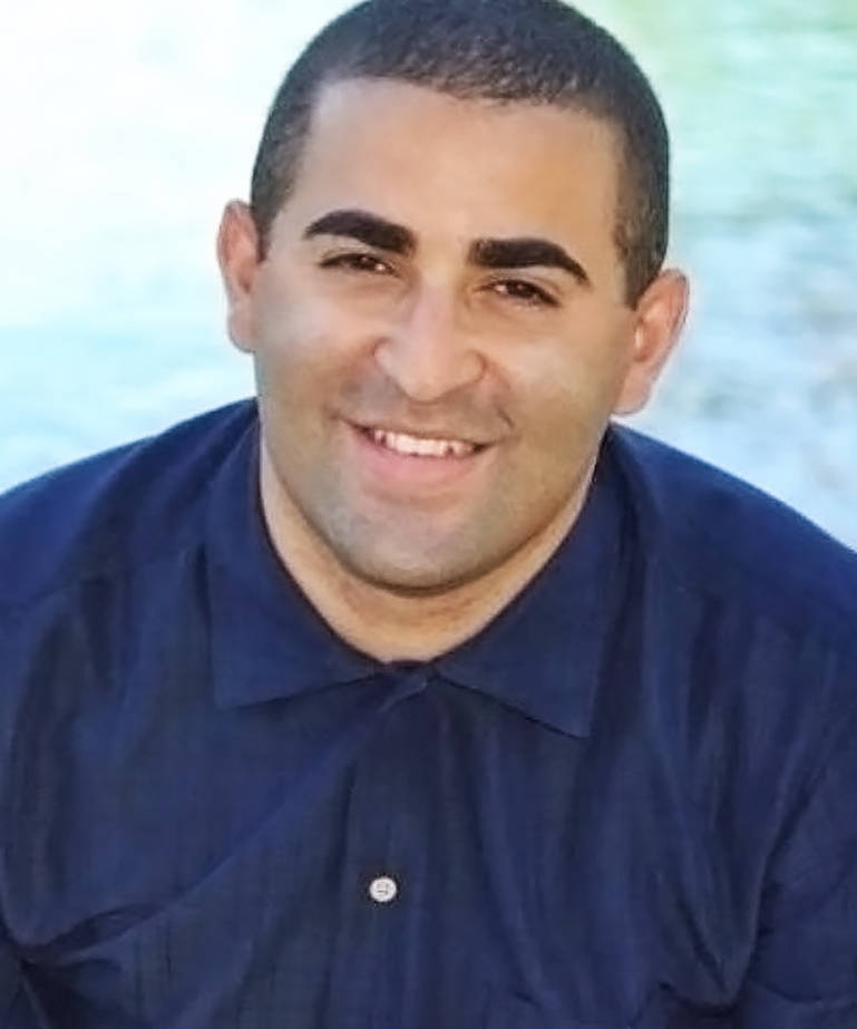 Dominican male wearing a blue buttoned down collared shirt. The image zoomed in to his head and shoulders. He sits in front of a body of water, smiles directly at the camera.