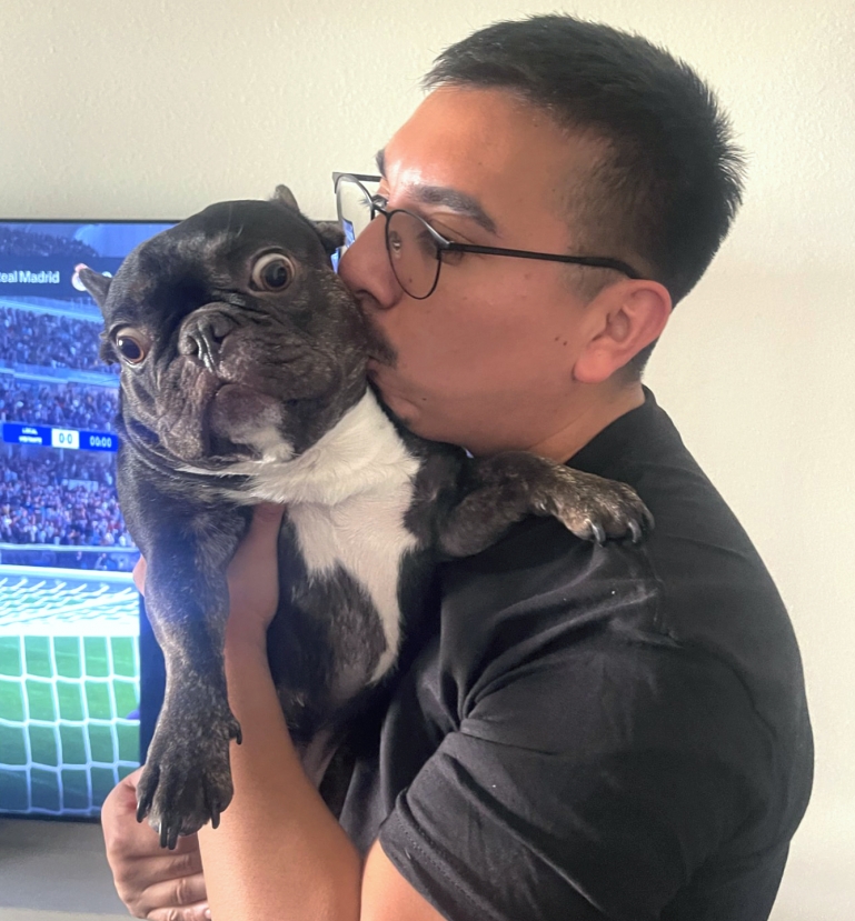 A photo of Jose lifting his grey French bulldog up and kissing it on the cheek.