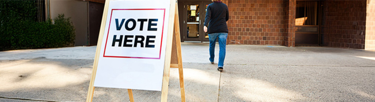 A brick building polling place with a stand that says, "Vote Here".