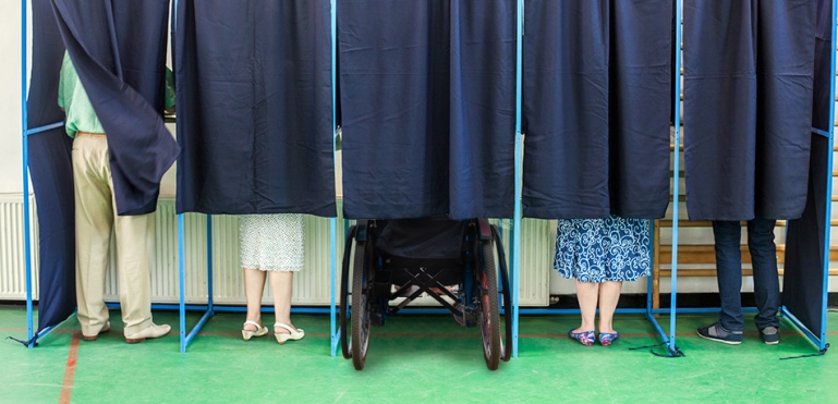 Image of legs of people behind a curtain at a voting boot. One of the voters is in a wheelchair