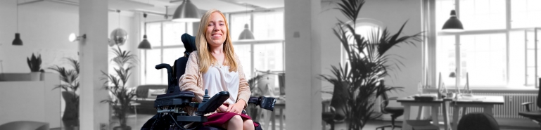 A women in a electric wheelchair working in a office. She is smiling.