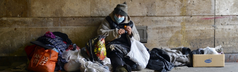 A homeless man in the streets with a face mask on.