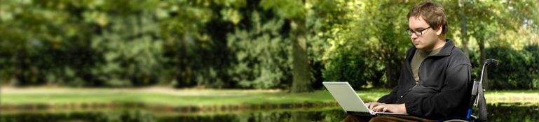 young disabled student learning at the park next to a pond while using his laptop