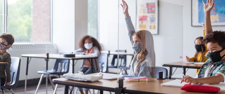 Several young children in a classroom. They are spaced apart and are wearing medical masks due to Covid-19.