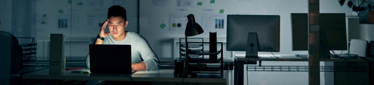 An employee working alone on his computer in a dark office. He has a worried look on his face.