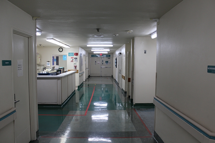 The hallway is lit with multiple fluorescent lights. The floors are shiny with red taped lines that depict where patients are allowed to walk. When occupied by patients, some may be found standing with their backs against the wall as Disability Rights California staff walked by.