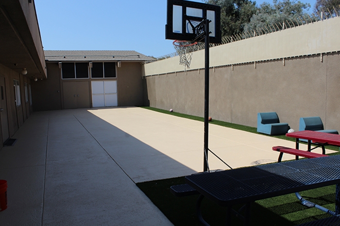 The patio has a basketball hoop and court which is a small strip of astroturf that separates the patio from a tall concrete wall topped with metal siding and spikes angled inwards towards the patio.