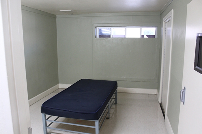 The room has one seclusion bed with a blue, synthetic mattress on top of a gray metal frame near the middle of the room. There is one small horizontal skinny window at the top of the wall opposite the entrance.