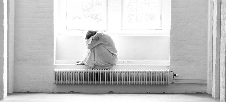 A photo of a women sitting alone by a window in a institution. Her face is hidden between her knees.