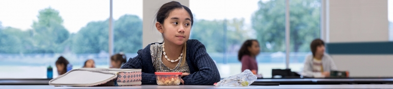 Lonely schoolgirl looks to the side while eating by herself in the school cafeteria. Other students are eating together in the background. 