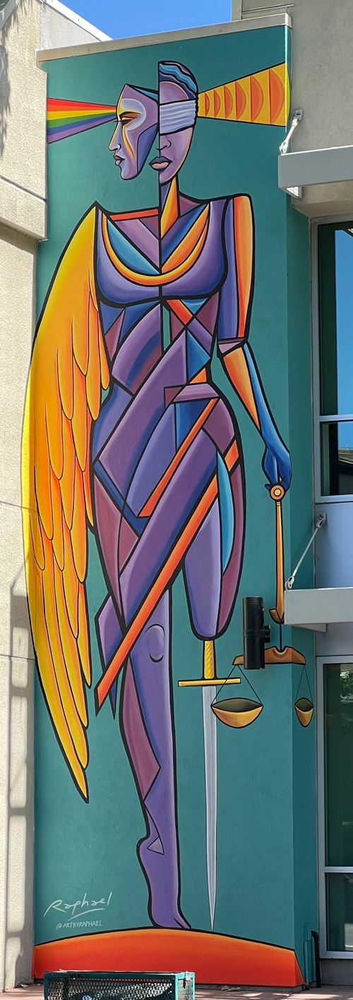 Photo of the showing a multi colored abstract mural  on the outside of DRC's building. She has a rainbow shining out of her right eye and wave forms thing out of her left ear. For her right arm she has a golden wing which is the length of her body. Her left leg has a sword for a prosthesis. Her left hand is holding golden scales.