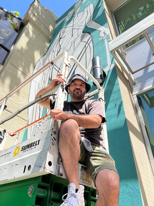 A photo of Raphael sitting on a lift with the mural behind him in progress.