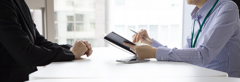 Cropped photo of two people at a desk showing only their arms and hands. One of the people is being interview by the other.