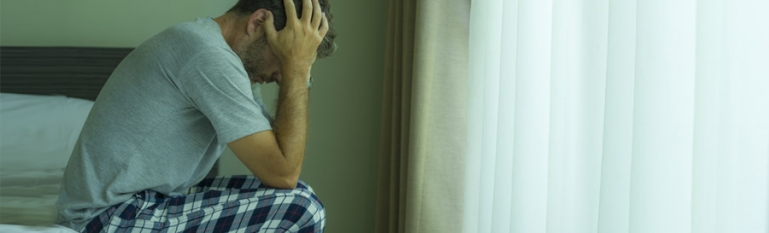 A man with mental health issues sits on the edge of a hospital bed with his head in his hands looking down.