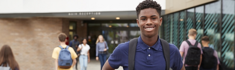 i young black student smiling in front of his school while other children enter.