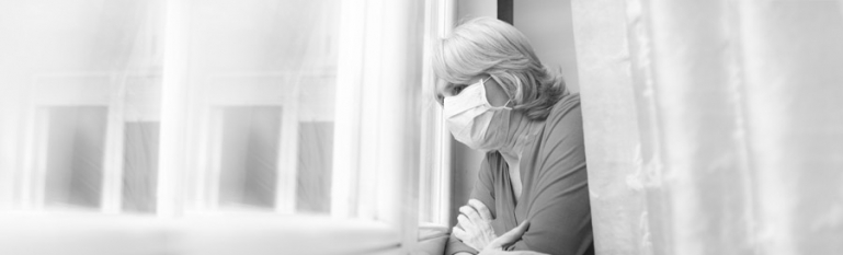An elderly woman wearing a medical mask looking out her apartment window.