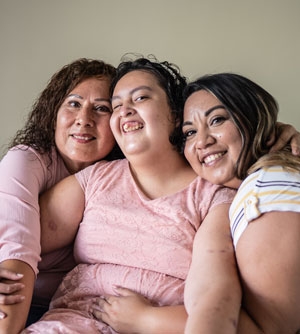 A Hispanic woman with a developmental disability smiling with her mother and her elder sister on either side of her.