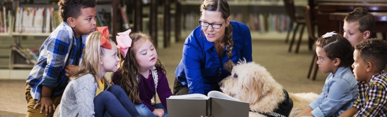 A teacher sitting down with her students reading a book. A few students have disabilities.