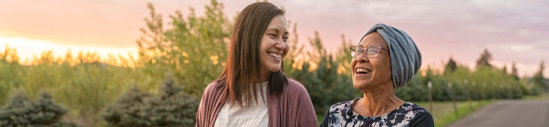Mixed race mother and daughter relaxing outdoors together. A senior woman with walks with her adult daughter at sunset down on rural road. The affectionate pair support each other while walking & talking with their arms linked.