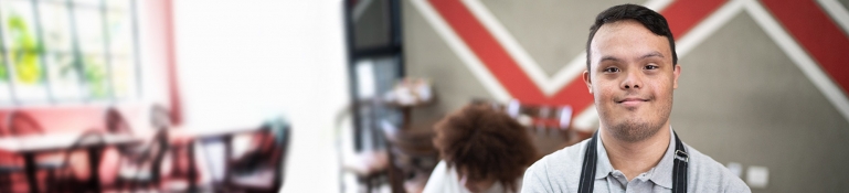 Portrait of a pridful special needs waiter standing with arms crossed in a coffee shop, with a customer in the background.