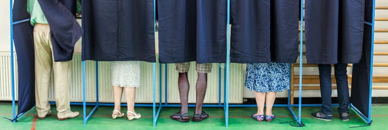 Photo of several voting booths. They each have a curtain covering the booths and only the voters legs and feet are showing as they vote.