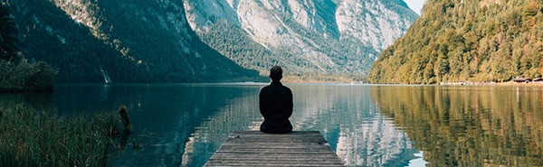 A lone person sitting on a boat dock overlooking a lake. There are mountains in the background. The image is calming.