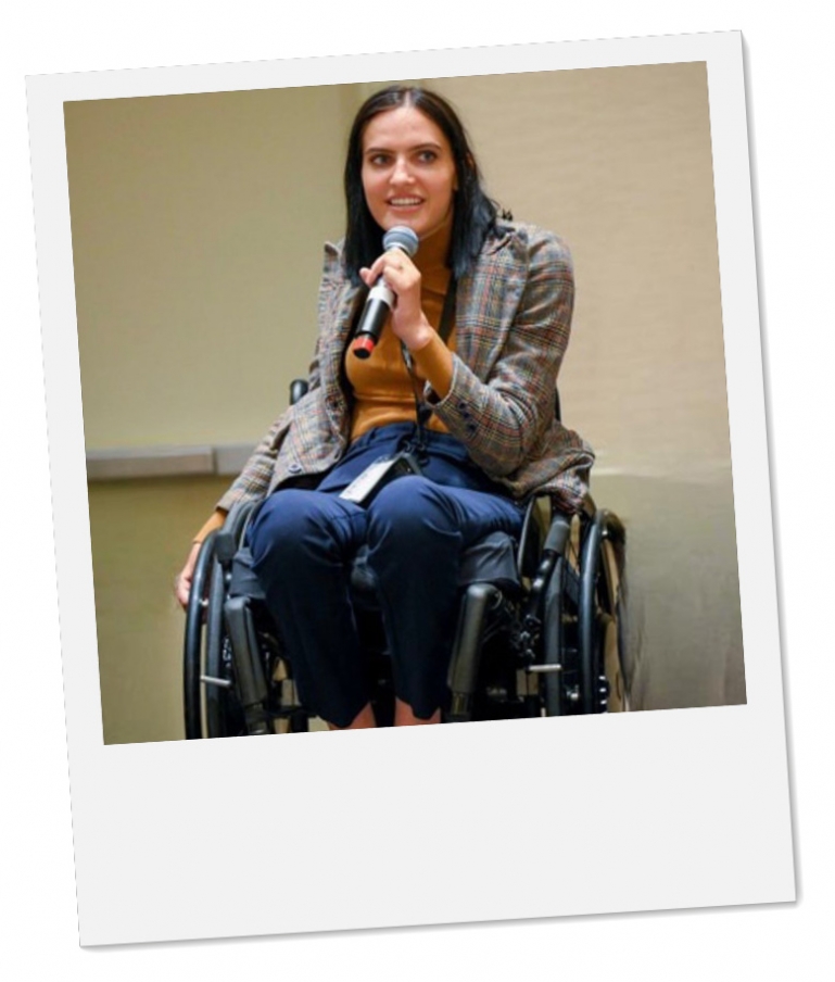 a white individual with dark brown hair sitting in a power-assist wheelchair, wearing a yellow, navy, and brown plaid suit and speaking into a handheld microphone.