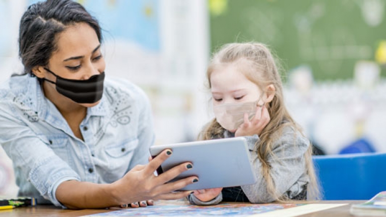 Image of a woman teaching a young girls who has a disability. They are both wearing COVID masks.