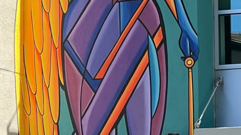 Photo of the showing a multi colored abstract mural  on the outside of DRC's building. she has a rainbow shining out of her right ey and wave forms thing out of her left ear. For her right arm she has a golden wing which is the length of her body. Her left leg has a sword for a prosthesis. Her left hand is holding golden scales.
