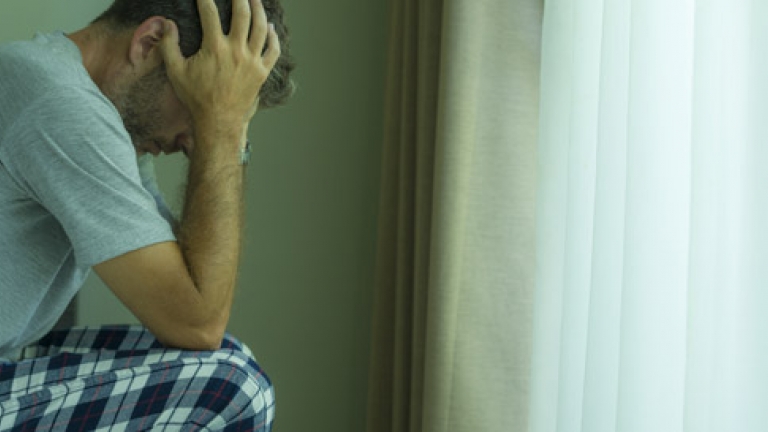 A man with mental health issues sits on the edge of a hospital bed with his head in his hands looking down.