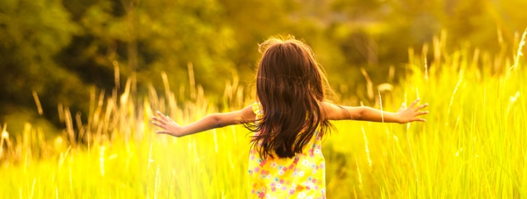 Photo of a young girl with a unknown disability with her arms out in a field. She has her back facing you as she is walking around happy.